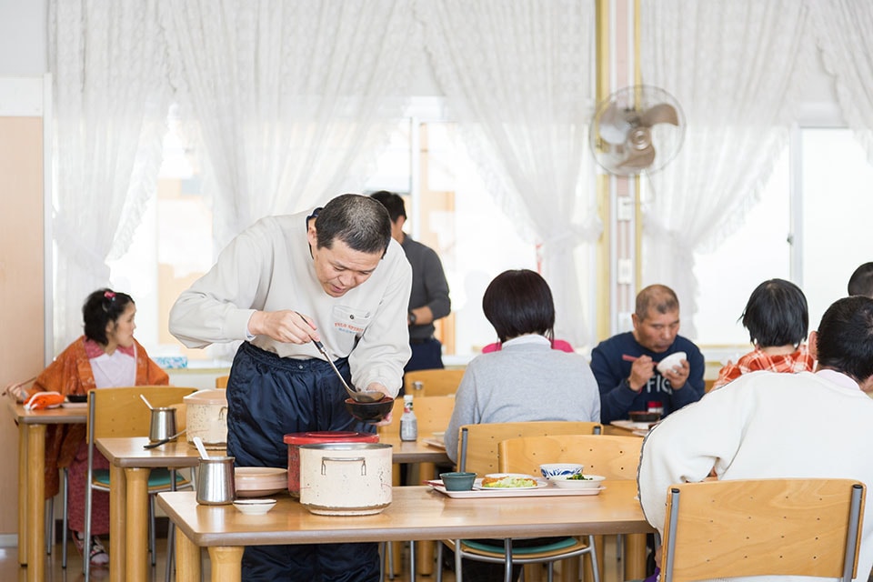 こうほうえんのしょくどうで、みやざきさんが、みぎてにおたまをもって、うつわにしるものをよそっているようす。