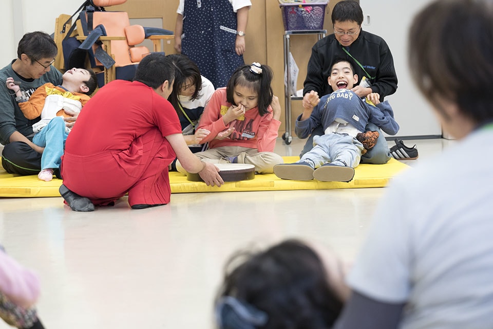 新井さんが太鼓などの打楽器を持って、円になって座っている子どもたちのところを順にまわる