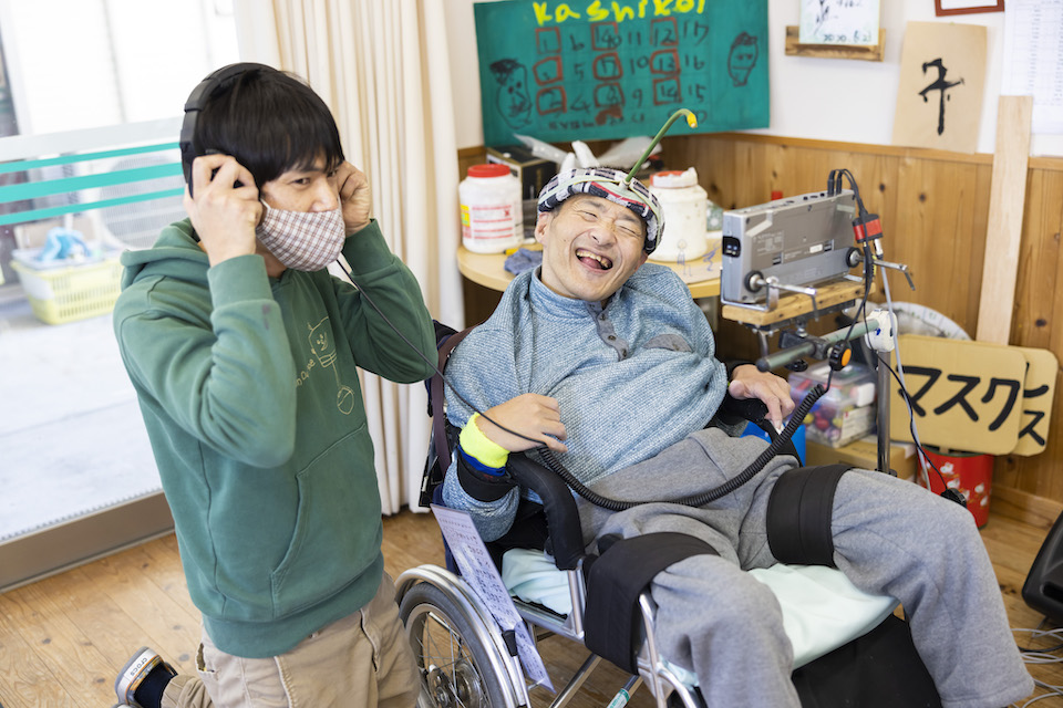 車椅子のすすきさんの横でヘッドフォンで音を確認する三上さん