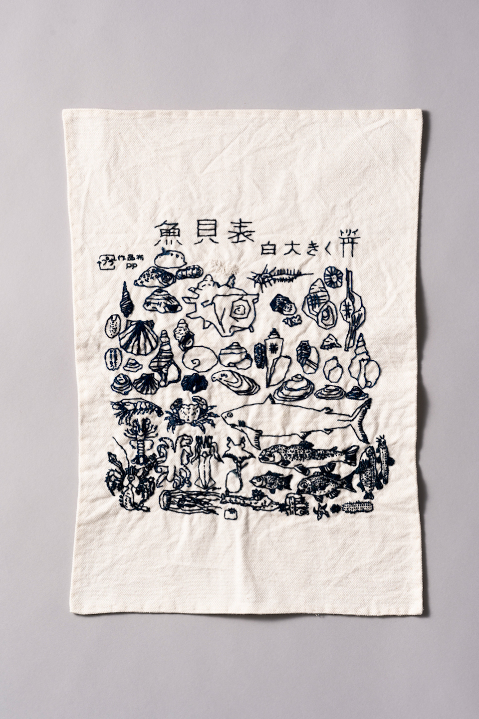 五十嵐朋之さんの作品画像。白い布に貝や魚が細かく刺繍で縫い付けられている。「貝魚表」という漢字が一番上方に塗ってある。