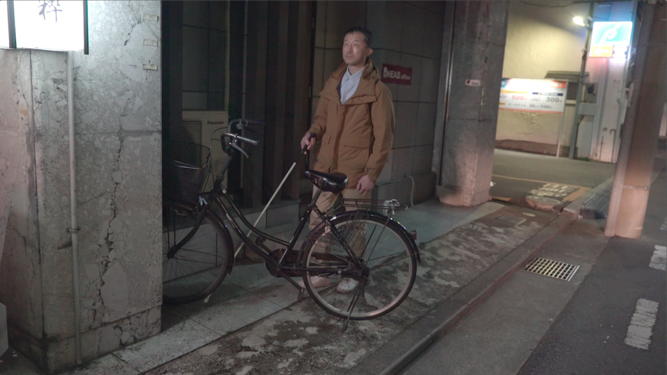白杖を手に、夜の歩道をひとり歩く白鳥さん。歩道にはみだして駐輪している自転車が行くてを阻む。