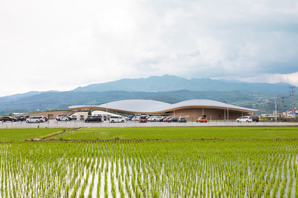 【写真】隣接する田園風景から臨む外観。背後の蔵王連峰に呼応した形状であることがわかる