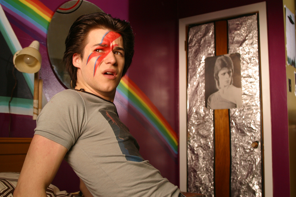 【写真】顔に赤い稲妻を描いた青年時代のザックが自室のベッドに寝そべり、驚いた表情で外を見つめる。ドアにはデビッド・ボウイのポスターが貼られる。