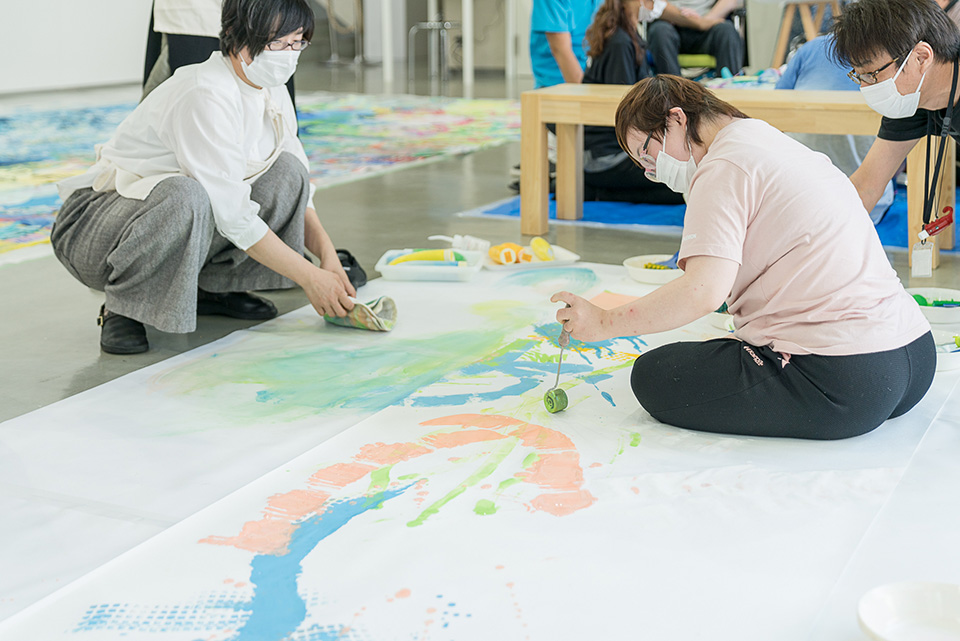 【写真】ワークショップで大きな紙の上でローラーで絵を描く参加者とそれを見守る二村さん