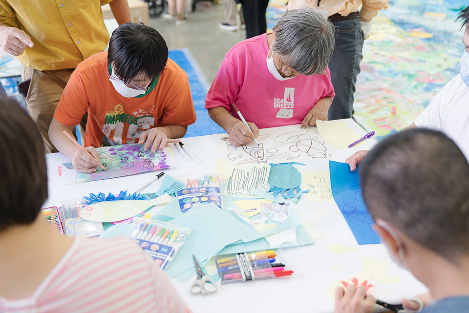 【写真】テーブルの上に色とりどりの紙とサインペン、3人の参加者が絵を描いている。