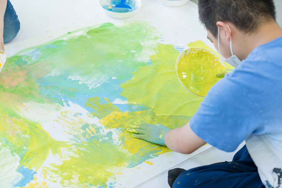 【写真】参加者のひとりが素手で絵の具を紙に塗り付ける。青、緑、黄色のカラフルな作品ができていく。
