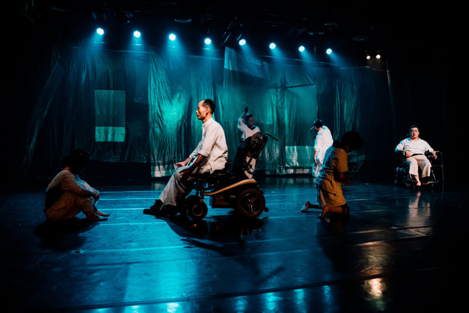 [Photograph]On stage, performers with various different disabilities are performing in a scene where each one is facing a different direction.