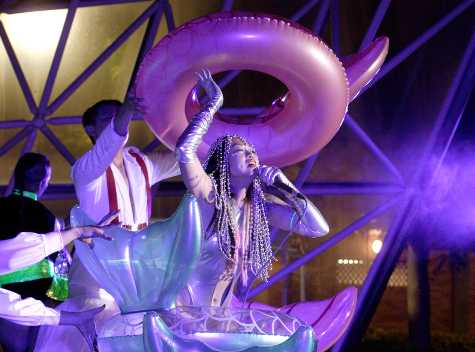【写真】人魚の姿をしたベティー・アップルが舞台上でマイクを持って歌っている。その周りにはダンサーたちが浮き輪を持っている