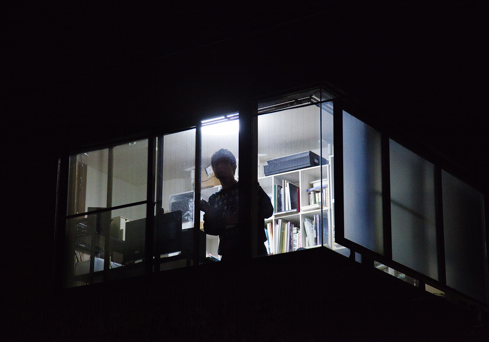 【写真】金川さんのシルエットが電気の灯った建物の窓辺から外を覗く様子