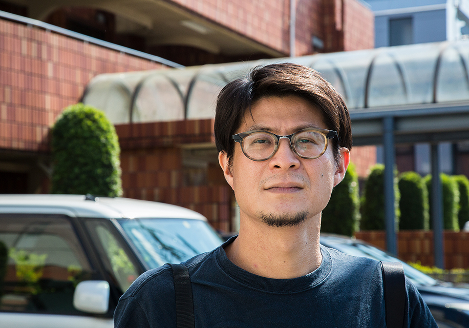 【写真】晴れた日中、マンション入り口付近の駐車場を背景に、金川さんが正面を向いているバストアップの写真