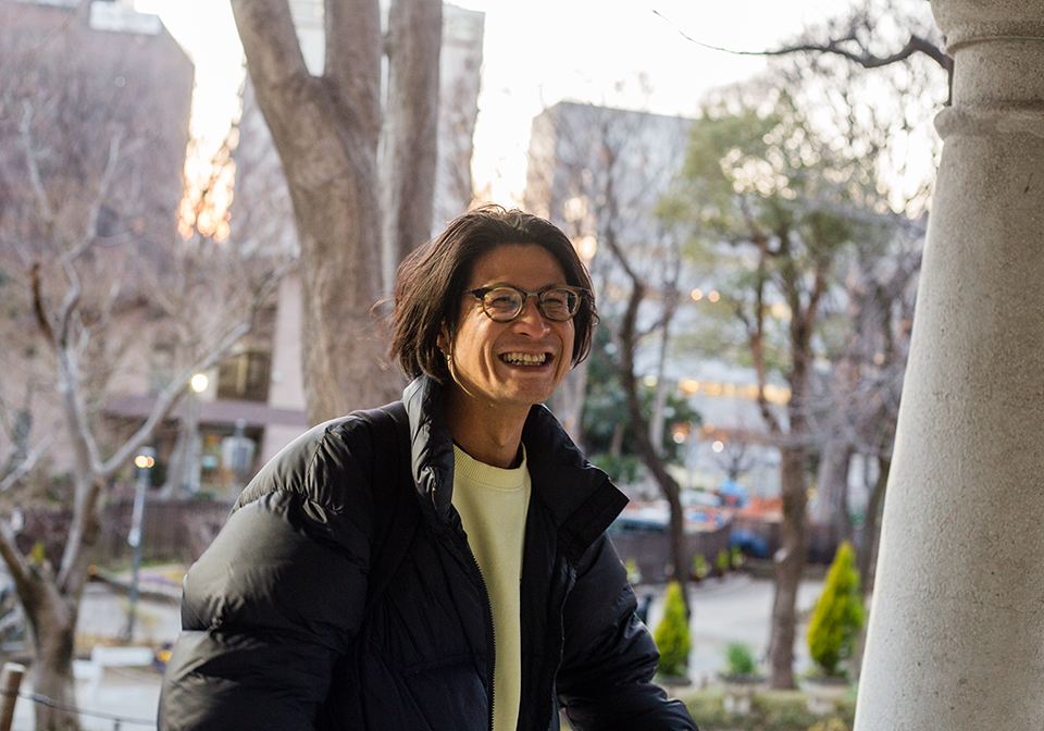 【写真】枯れ枝の木々が並ぶ公園を背景に、黒いダウンジャケットを着た金川さんが、笑顔で右正面を向いている様子