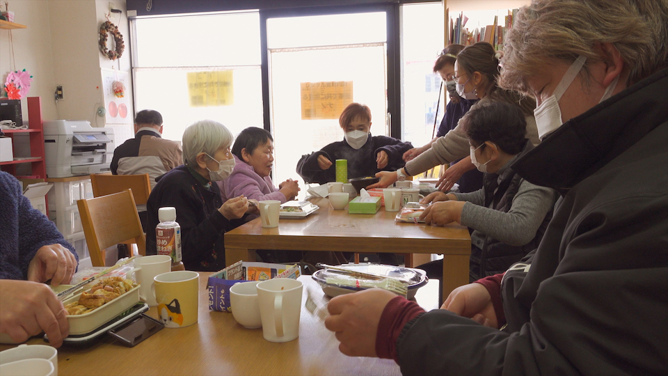【写真】地域で共に生きるナノの昼食風景。机の前には9人の人たち。それぞれ持参したり買ったりしたお弁当を広げて食べようとしている。