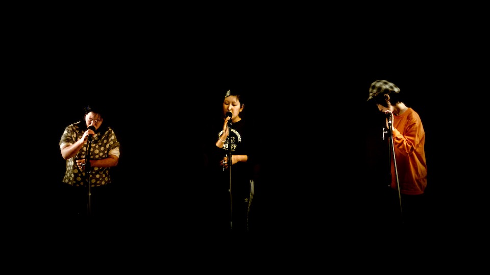 【写真】暗いステージの上。マイクの前に立って歌う3人のメンバーにスポットライトが当たっている。