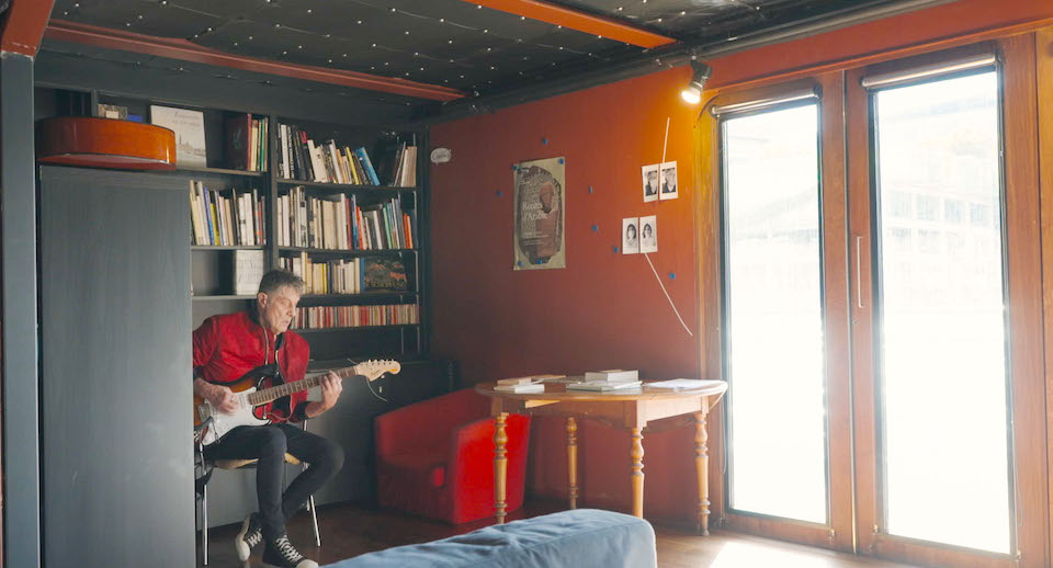 【写真】部屋の片隅、本棚の前でエレクトリックギターを弾き、歌をうたっている人がいる。赤いジャケットを着ている。大きな窓からは光が差し込んでいる。