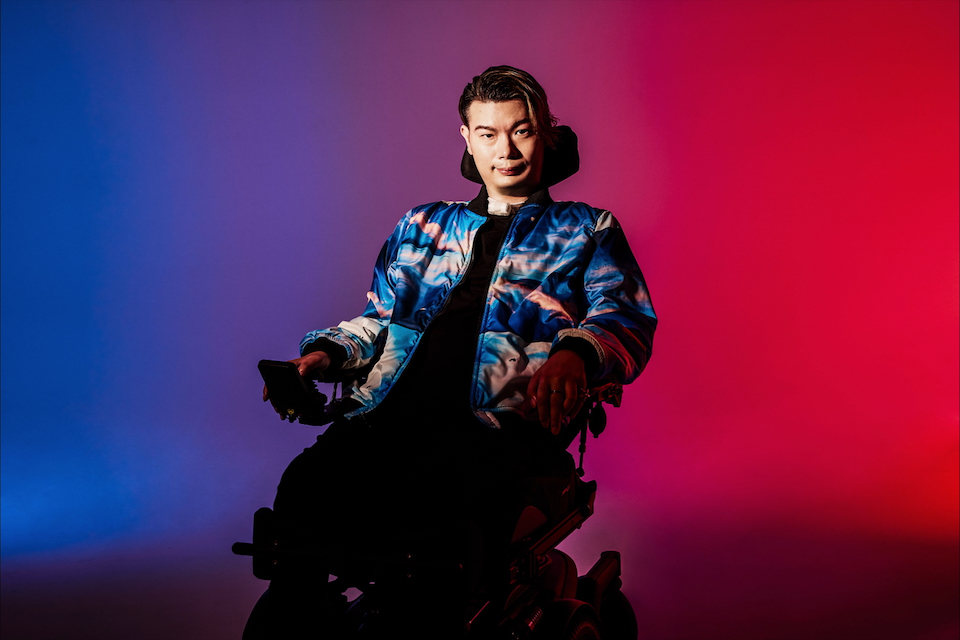 【写真】車椅子に座った武藤さんがこちらを鋭い眼差しで見詰めている。武藤さんは青と白とピンクの色がマーブル模様になったジャケットを着ている。背景は左側は青、右は赤の色がついており、とても鮮やかな色の写真になっている。