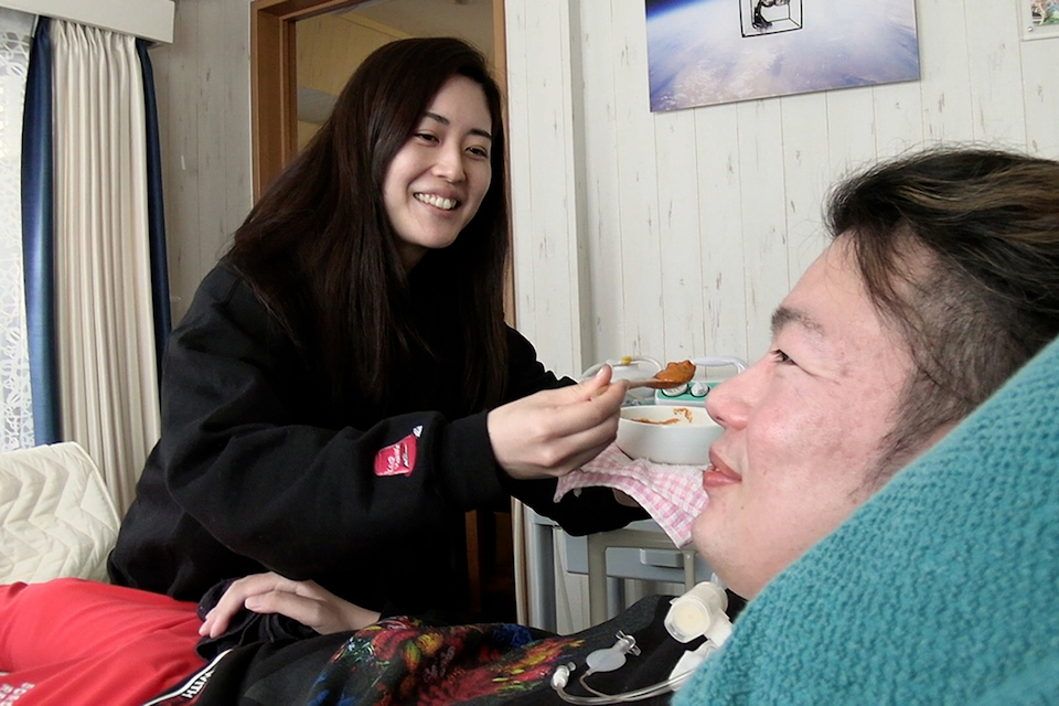 【写真】妻の木綿子さんが、座っている武藤さんの口元に食事を運んでいる