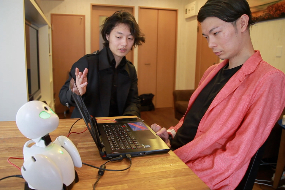 【写真】武藤さんと吉藤オリィさんが、パソコンを見ながら話している。パソコンの傍らには分身ロボットのオリヒメが置いてある。