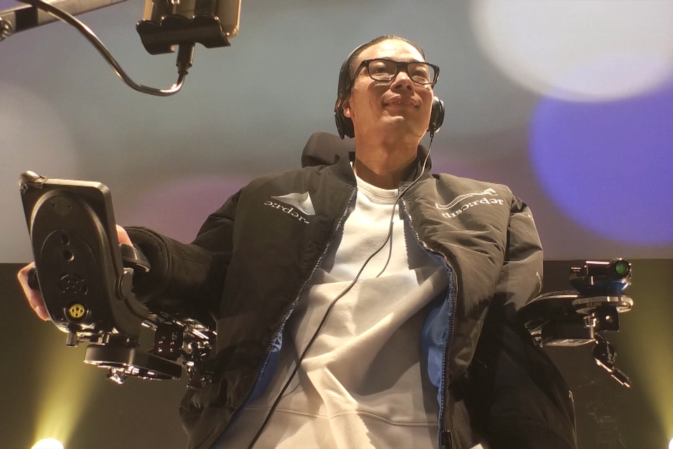 【写真】車椅子に乗り、黒いジャケットに黒縁メガネをつけた武藤さんが微笑んでいる。