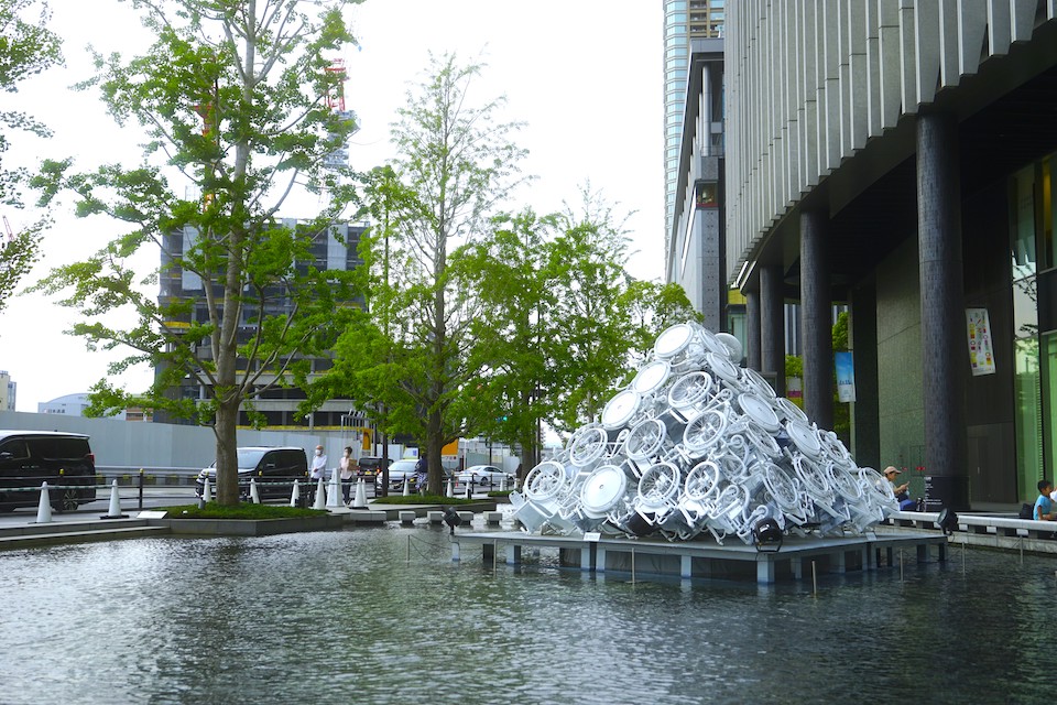 【写真】檜皮一彦さんの作品。ビルの横にある人工池の上に、白く塗られた車椅子が三角錐の形に積み上げられている。