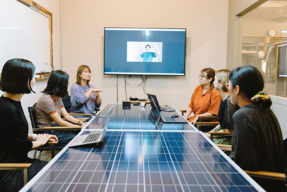 【写真】女性6名が壁にかけられたモニターと机を見ながら手話で会話している。モニターでは手話をしている男性が写っている。