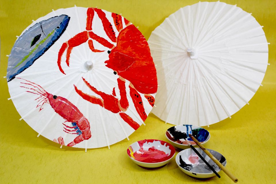 白い和傘が2つ。左には金沢らしくカニやエビ、魚が描かれている。手前に絵具と筆がおかれている。