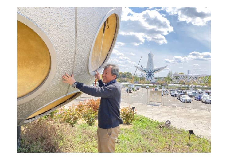 【写真】ひろせこうじろうさんが、野外彫刻をさわっている様子。遠くに太陽の塔がみえる。