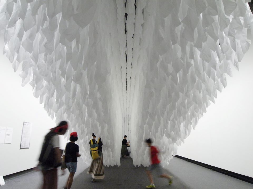 【写真】展示空間内の天井から白い布のような作品が垂れ下がっている。下に鑑賞する人々。