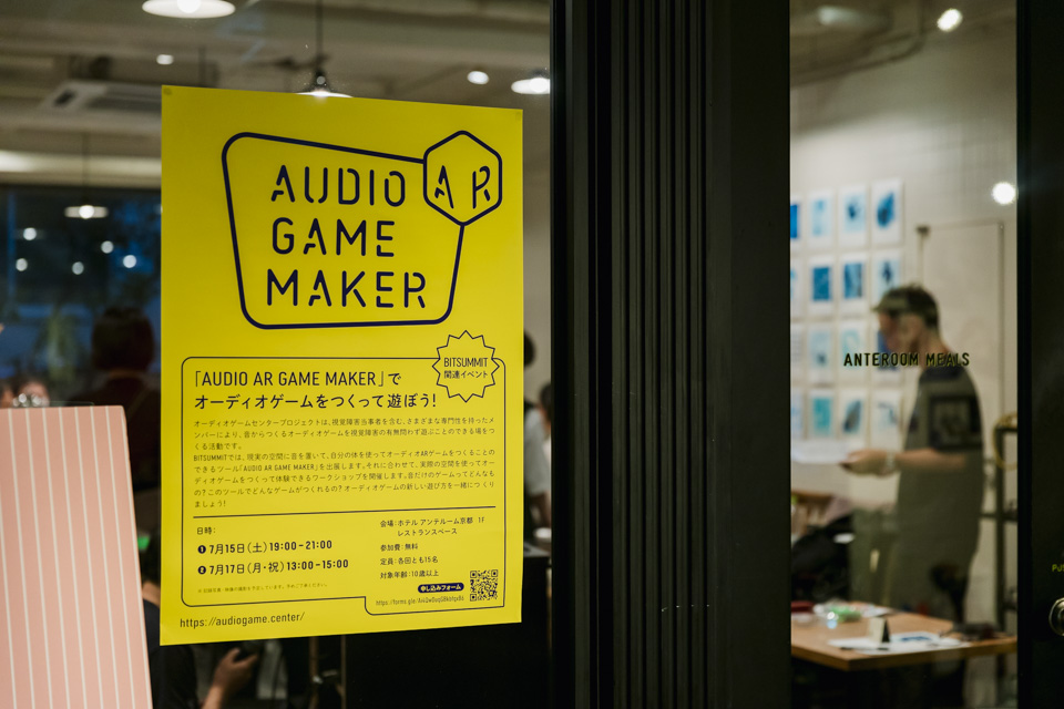 【写真】扉に貼られたワークショップのチラシ。タイトルに、「AUDIO AR GAME MAKER」でオーディオゲームをつくって遊ぼう！、とある。扉の奥ではワークショップの準備が始まっている。