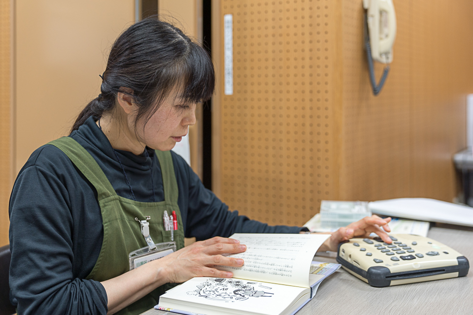 【写真】椎原さんがテーブルの前に座り、本を開きながら、プレストークを操作している。本には文章と挿絵がある。
