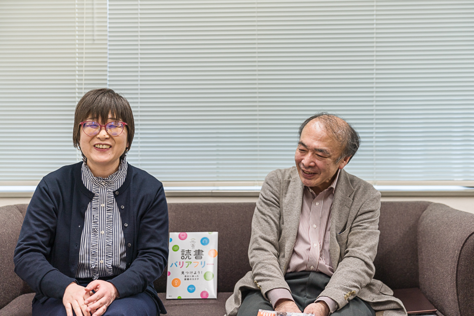 【写真】笑顔でソファーに座る二人。向かって左が村上さん、ショーットカットで赤いメガネ、青いストライプのシャツに紺色のカーディガンを羽織っている。右に笑顔の成松さん。間に読書バリアフリーの本。