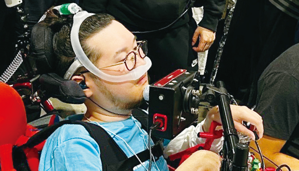 【写真】車椅子に座ったJeniさんが自作したコントローラーを顎で操作している