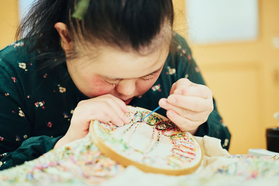 【写真】メインビジュアルと同じ写真。綾さんが丸い木枠の型をはめた布を顔に近づけ、黙々と刺繍に取り組む様子。