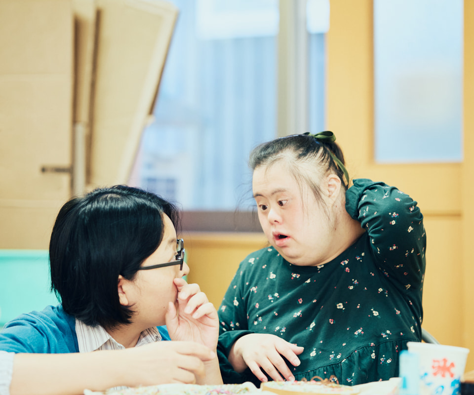 【写真】アトリエ内で上遠野さんと綾さんが向かい合って話している。上遠野さんはしゃがんでテーブルに腕を置き、席につく綾さんと目線を合わせている。