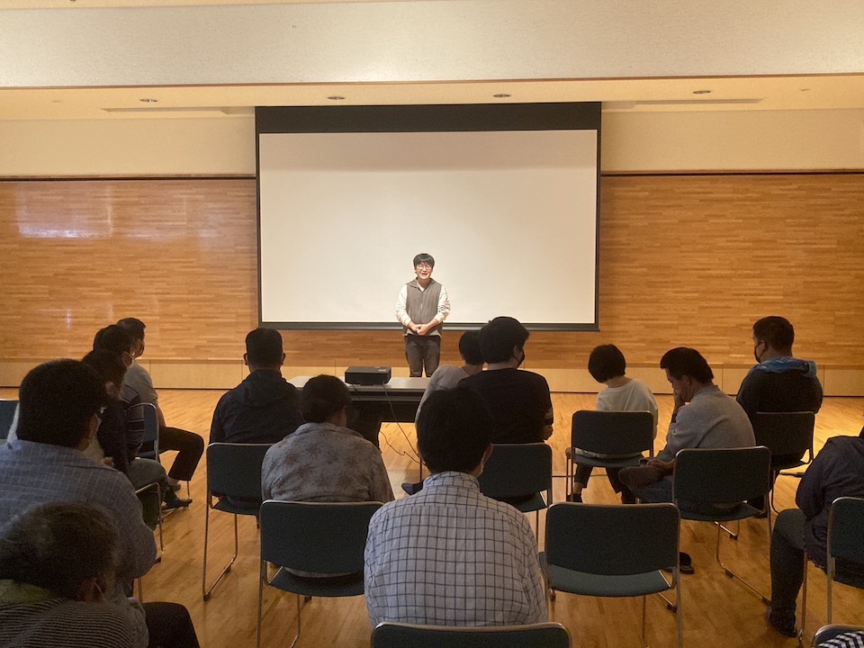 【写真】関係者試写会のスクリーン前で挨拶をする映画監督の青柳さん。20人ほどの鑑賞者が椅子に座って話を聞いている様子。