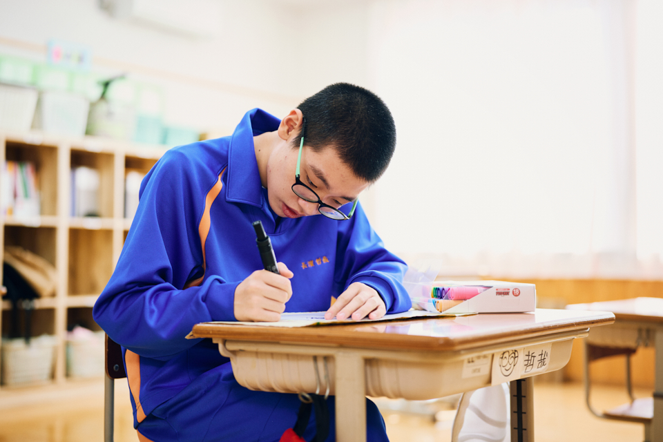 【写真】長濱哲哉さんが教室の机に向かい、油性ペンで絵を描いている様子。
