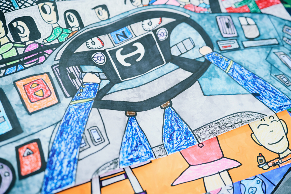 【写真】車を運転する様子を描いた絵。運転手の目線から見た構図になっており、ハンドルを握る手や自身の脚が描かれている。ハンドルのまわりにはスイッチやメーターがあらゆるところに付いており、フロントウィンドウの先には、道行く人の姿がある。