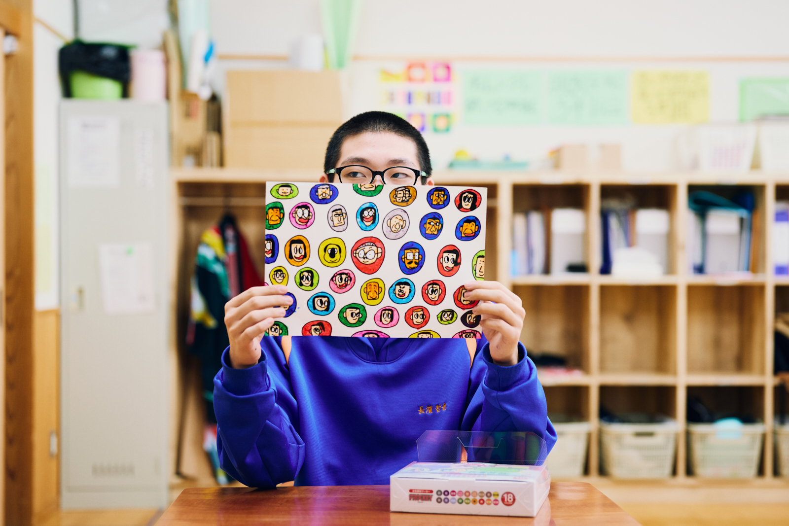 【写真】山形県立村山支援学校本校教室内にある机につき、両手で持った絵の作品から顔を覗かせる、生徒の長濱哲哉さん。