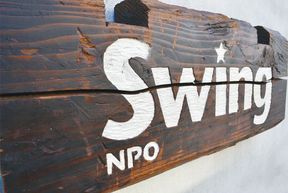 【写真】NPO法人「スウィング」の看板。木目が浮き出た木製看板に、白いゴシック体の文字でNPO Swingと書かれている。