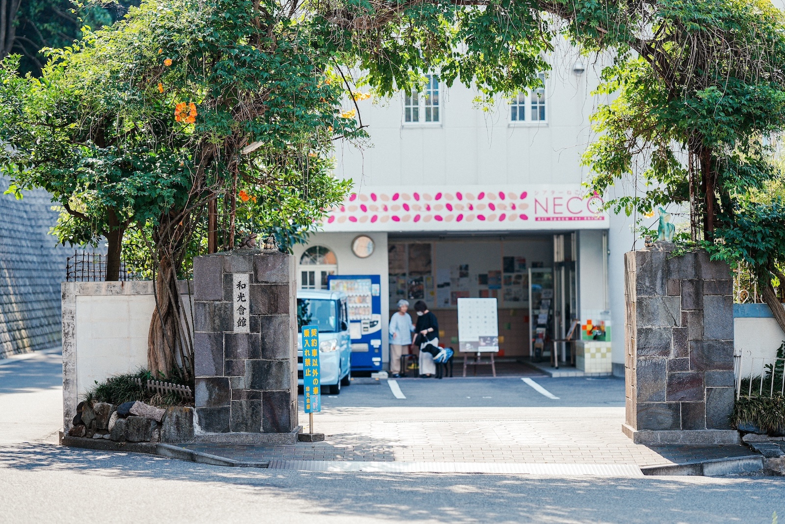 【写真】愛媛県・松山の〈和光会館〉外観。石造りの門の両脇に木が植えられ、地面に木漏れ日が映っている。門の先には駐車場と、同館に併設された、NPO法人シアターネットワークえひめが運営する〈シアターねこ〉の玄関口がある。