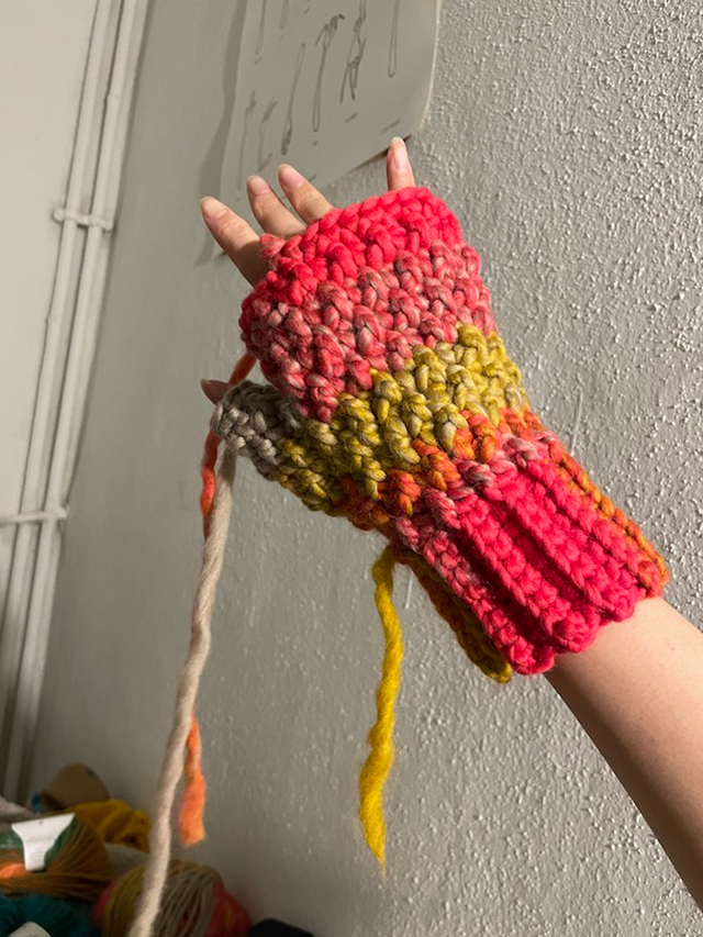 【写真】編みかけの手袋を手にはめている様子。赤や黄、オレンジ、薄ベージュの糸が編まれ、きれいな模様をつくっている。