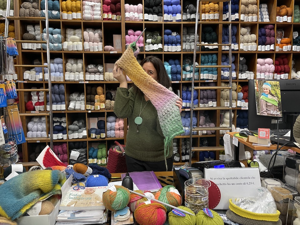 【写真】女性が編みかけのマフラーを持っている様子。マフラーの色はベージュから黄、オレンジ、ピンク、白、ライムグリーンへと徐々に変わっていくグラデーションになっている。