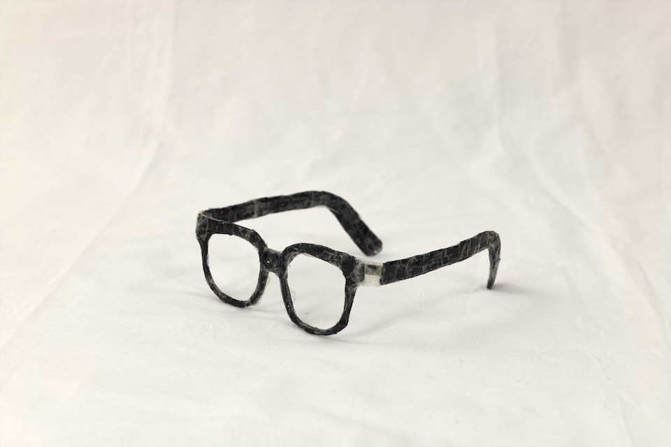 【作品画像】白い背景に1つの眼鏡が置かれている。遠目に見ると太い黒縁の通常のメガネに見えるが、よく見ると少し歪んでいるのがわかる。
